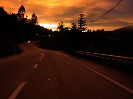 Carretera de dos carriles al anochecer, con un coche que se aproxima a lo lejos con las luces encendidas.